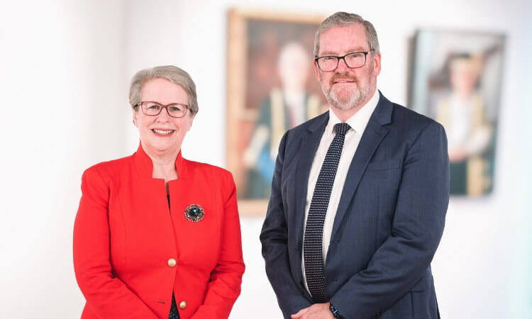 Two individuals, one in a red blazer and the other in a dark suit, pose side by side in an art gallery with framed portraits in the background.