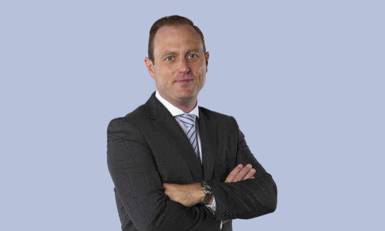 A man in a suit and tie stands with his arms crossed against a plain light blue background.