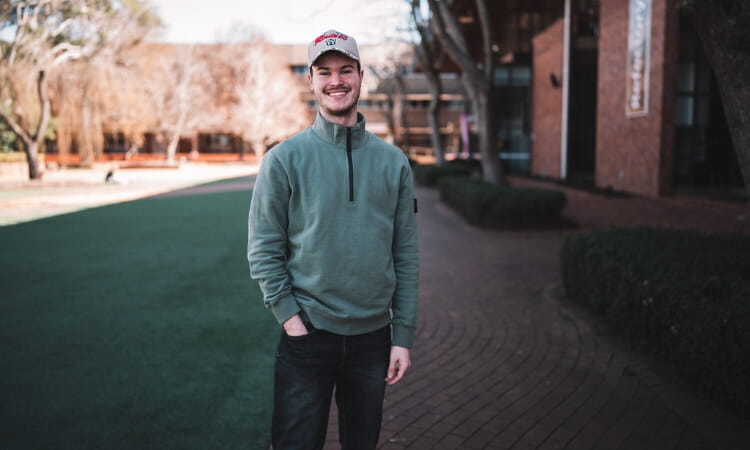 A man in a green pullover and jeans smiles while standing on a brick pathway with a lawn and buildings in the background.