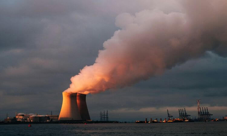 A nuclear power plant emits steam from cooling towers at sunset, set against a cloudy sky and located near a body of water with industrial structures in the background.