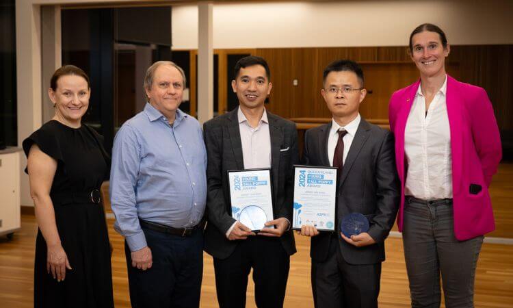 Five people stand indoors, with two individuals in the center holding glass awards and certificates. Everyone is dressed in business or semi-formal attire, and they smile at the camera.