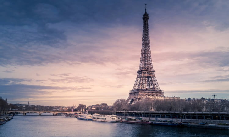 The Eiffel Tower at sunset with the Seine River in the foreground and boats docked along the riverbank.