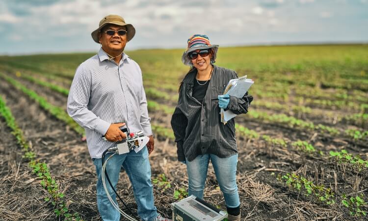 Two people stand in an agricultural field holding scientific equipment and notes, conducting research.