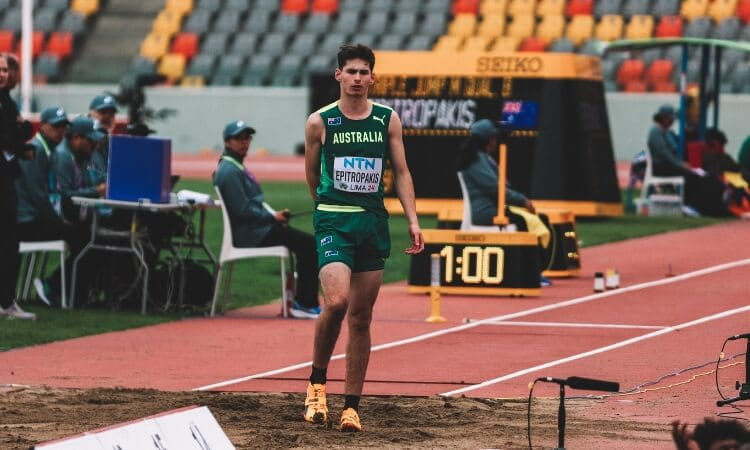 An athlete in a green uniform and bright orange shoes, representing Australia, stands on a track field with officials seated nearby.