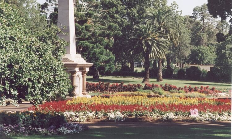 A garden with vibrant flower beds, a white monument on the left, lush green trees, and a palm tree in the background on a sunny day.