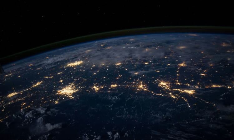 A view of Earth from space at night, showing brightly lit urban areas and a dark expanse of ocean and land.