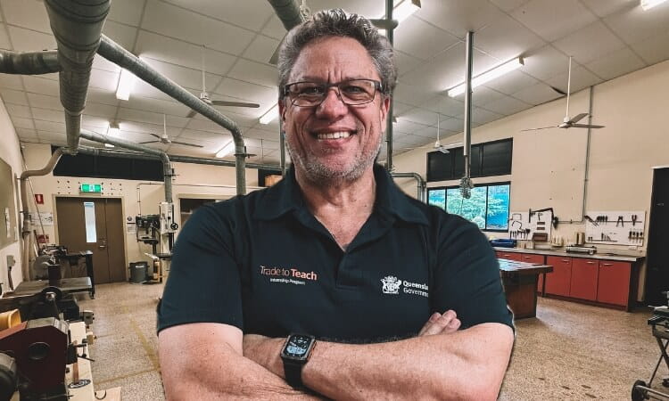 A smiling man with glasses and stands in a workshop with his arms crossed. 