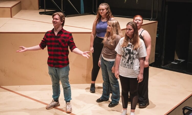 A group of five people stand on a stage, with one person in front gesturing widely while the others look on. 