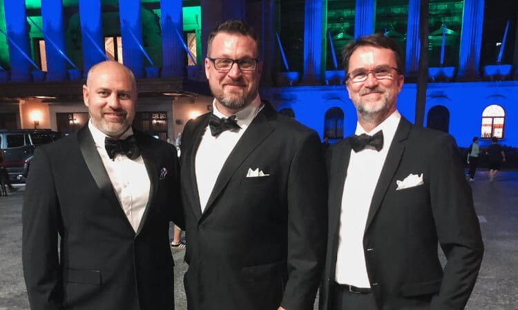 Three men in black tuxedos and bow ties stand together outdoors at night in front of a building illuminated with blue and green lights.