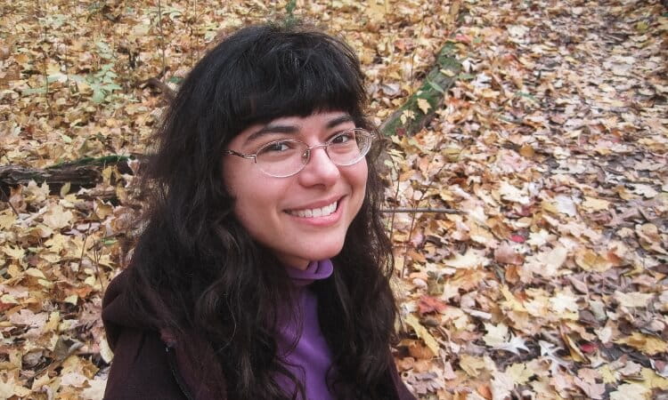 A person with long dark hair and glasses smiles while standing among fallen leaves in a forested area.