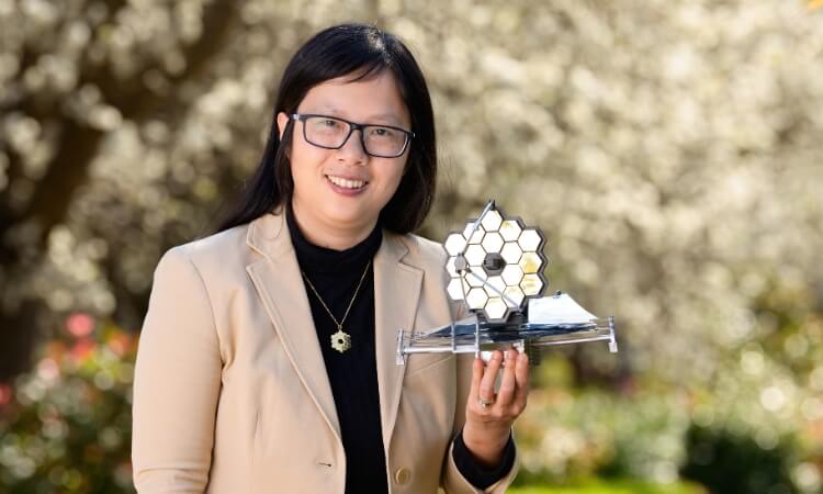 A person wearing glasses and a beige blazer holds a small model of a structure with hexagonal elements while standing outdoors.