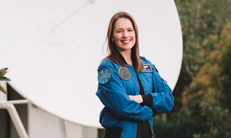 A woman in a blue NASA jacket stands with arms crossed, smiling, in front of a large satellite dish.