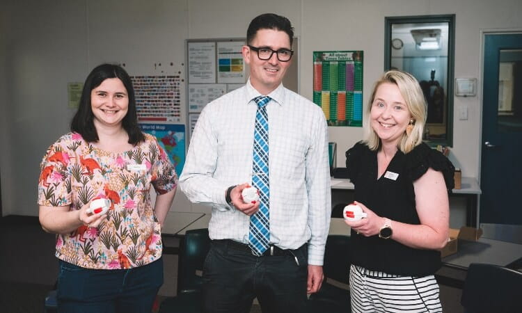 Three people, two women and one man, standing indoors and holding red and white objects while smiling at the camera. The background includes office or classroom elements.