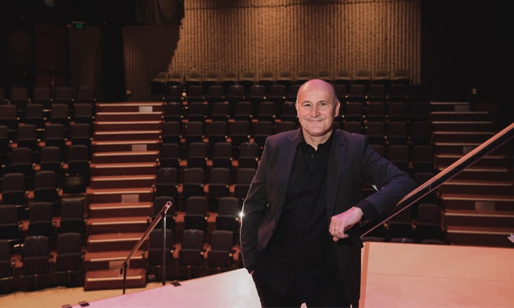 A person in a black suit stands on the stage of an empty auditorium, smiling at the camera.