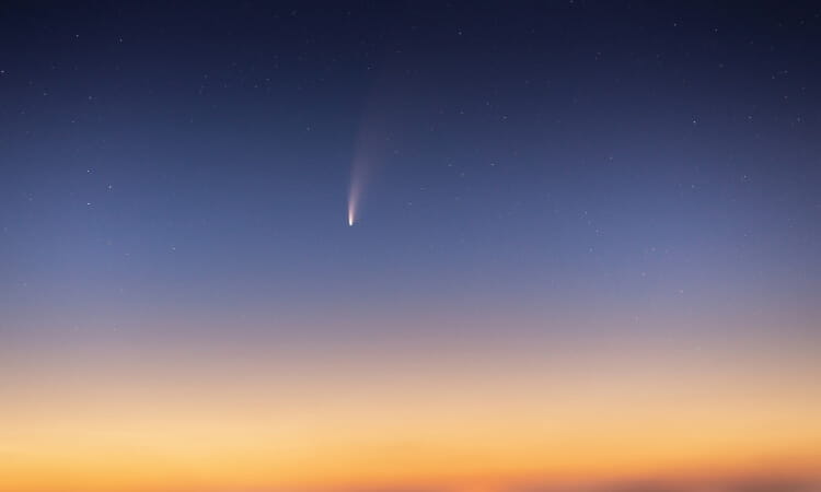 A comet with a visible tail streaks through the twilight sky, set against a gradient background transitioning from orange to deep blue.