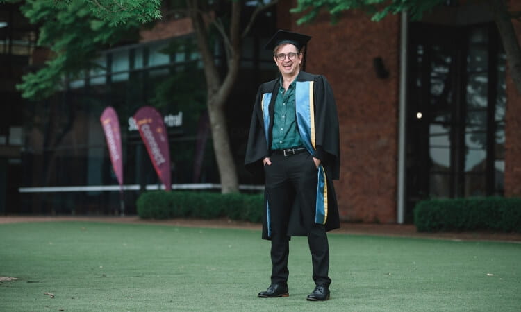 A person in graduation attire stands on a lawn in front of a building. 