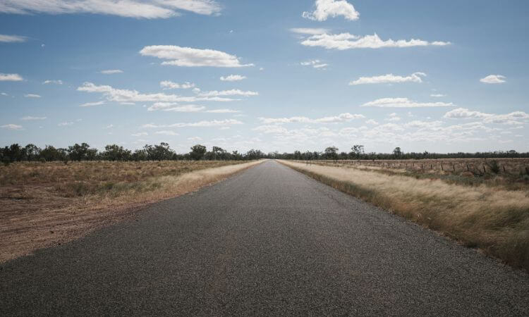 A long, straight road stretches into the distance under a blue sky with scattered clouds, flanked by grass and sparse trees.