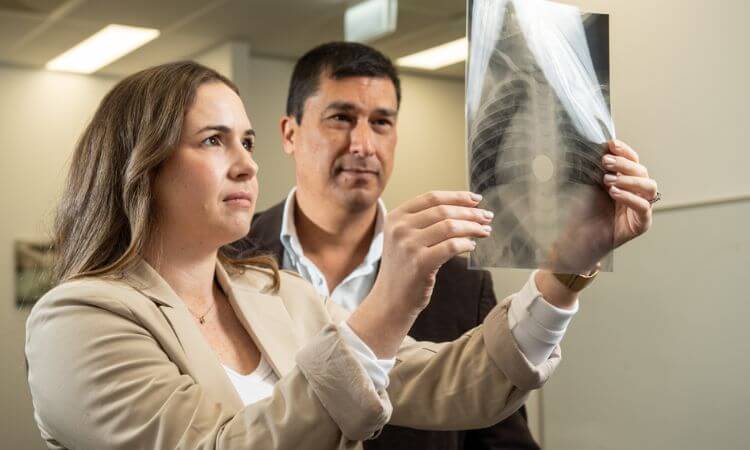 Two people examining a chest X-ray in a well-lit room. The woman holds the X-ray up while the man observes. Both appear focused and attentive.