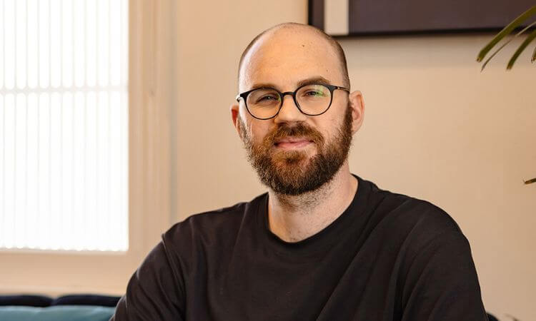 A man with a beard and glasses, wearing a black shirt, sits indoors in front of a light-colored wall.