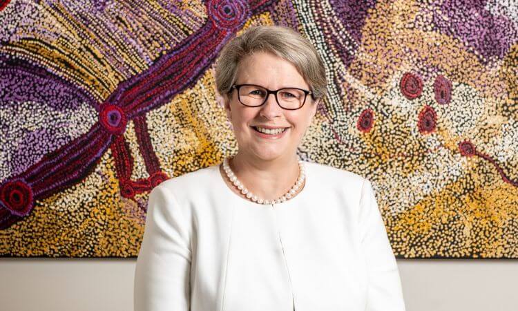 A person wearing glasses and a white outfit is smiling in front of a colorful abstract painting.