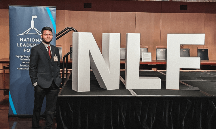 A man stands beside large letters "NLF" and a banner reading "National Leadership Forum" indoors.