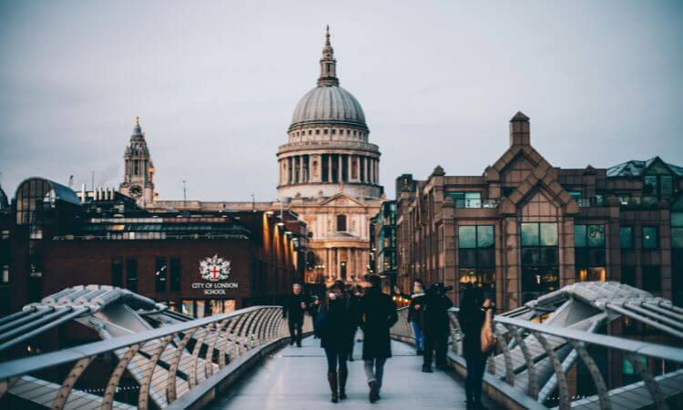 People walking on the Millennium Bridge in London with St. Paul’s Cathedral in the background under a cloudy sky.