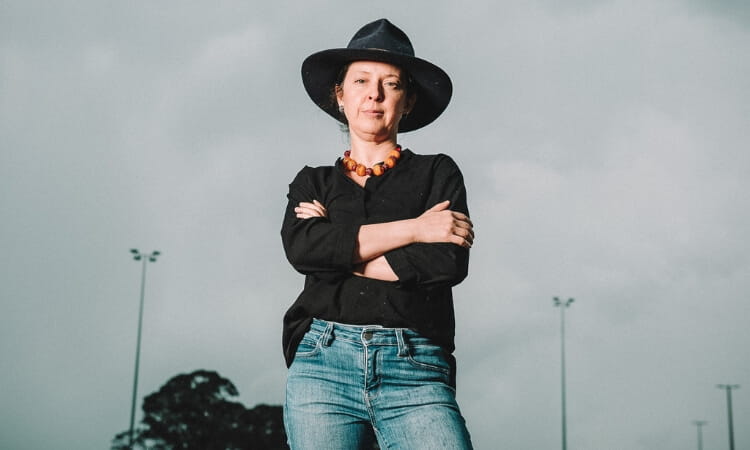 Person wearing a black hat, black shirt, and jeans stands outdoors with arms crossed, looking at the camera.