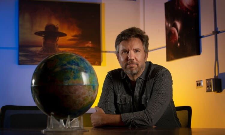 A man sits at a table with a globe in the foreground and space-themed art in the background.