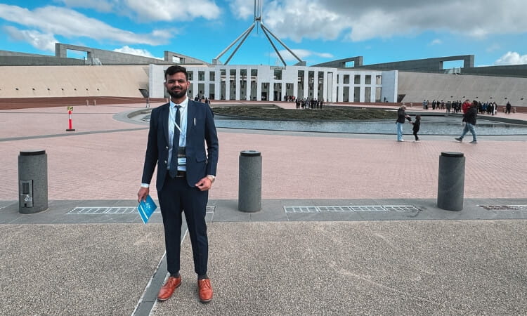 Person in a suit stands in front of a large building with a distinctive roof structure and flagpole, under a cloudy sky.