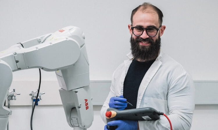 A man in a lab coat operates a large robotic arm using a handheld control device, standing in a laboratory setting.