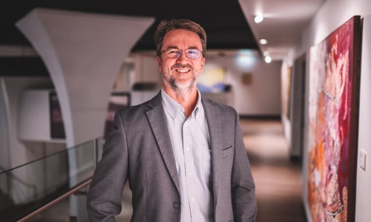 A man in a gray suit and glasses stands smiling in a modern hallway with art on the wall.