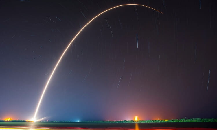 A long-exposure photo shows the arc of a rocket launch at night, with a bright trail against a dark sky and distant lights on the horizon.