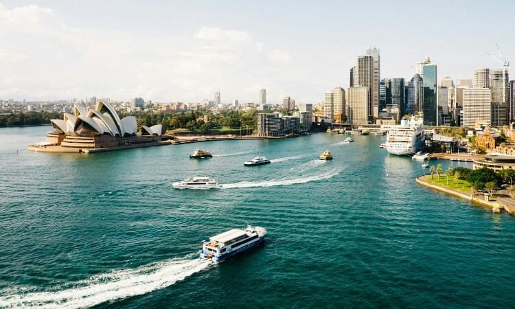 Aerial view of Sydney Harbour with boats on the water, the Sydney Opera House on the left, and city skyscrapers in the background.