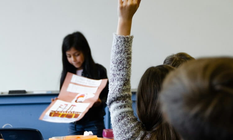 A student presents a project while another student raises their hand in a classroom.