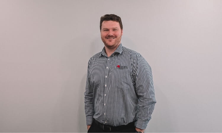 A person wearing a checkered shirt stands in front of a plain light-colored wall, smiling at the camera.