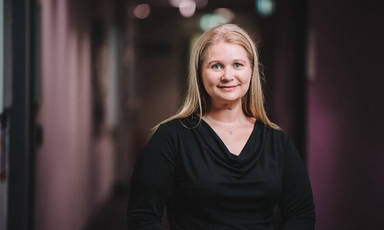 A woman with long blonde hair and a black top stands in a hallway with soft lighting.