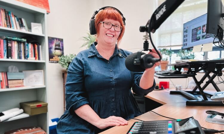 A woman with red hair, wearing headphones and a blue dress, sits at a desk with a microphone and audio equipment. 