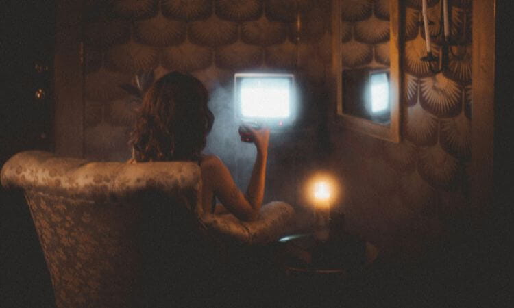 A person sitting in an armchair watches a small TV in a dimly lit room with patterned walls. A candle glows nearby.