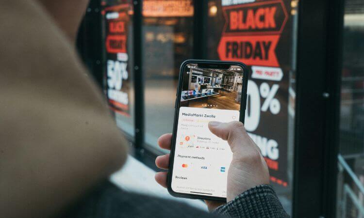 Person holding a smartphone displaying a store location app, standing in front of a shop window with Black Friday sale signs.