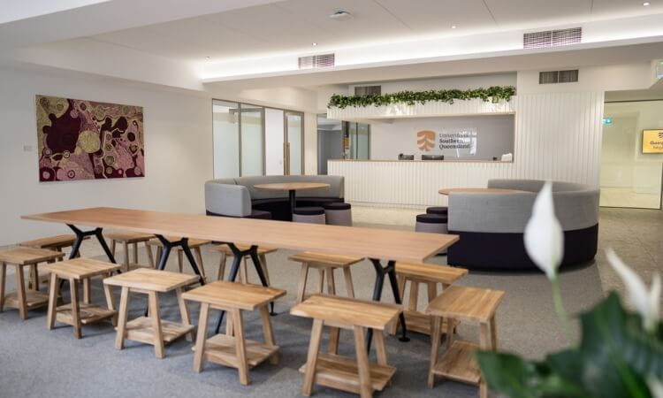 Spacious modern office lobby with wooden tables, stools, and a reception desk. Artwork and greenery adorn the walls.