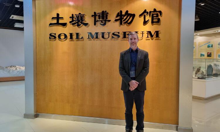 A person stands smiling in front of the Soil Museum sign on a wooden wall indoors.