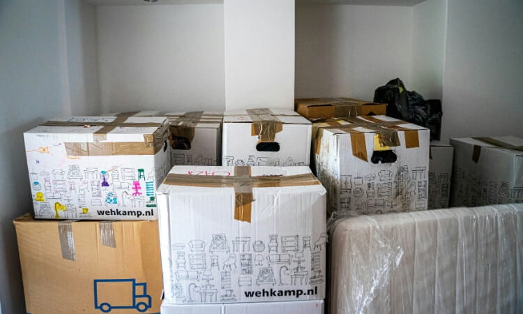 Several sealed cardboard boxes stacked against a wall in a room, with a wrapped mattress upright beside them.