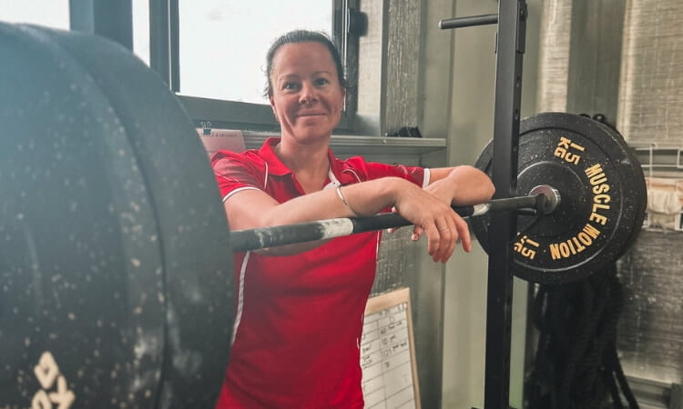 A person in a red shirt leans on a barbell with weights in a gym.