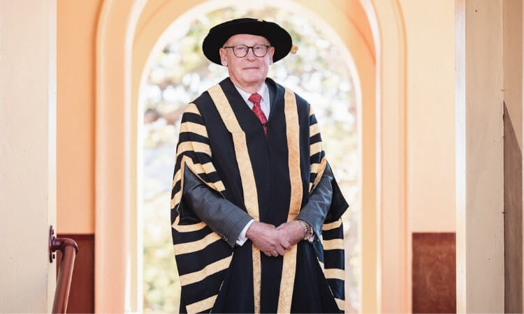 A person wearing academic regalia, including a black and gold gown and a cap, standing in an arched hallway.