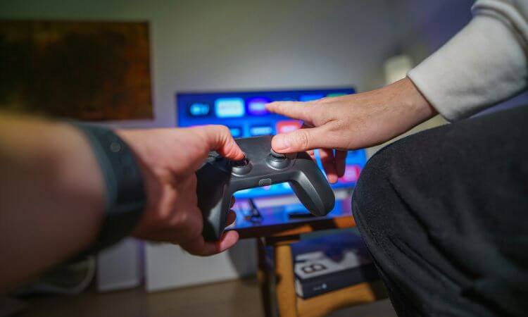 Hands holding a video game controller in front of a TV with a selection of apps displayed.