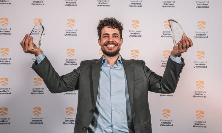 Person in a suit smiling and holding two awards in front of a University of Southern Queensland backdrop.