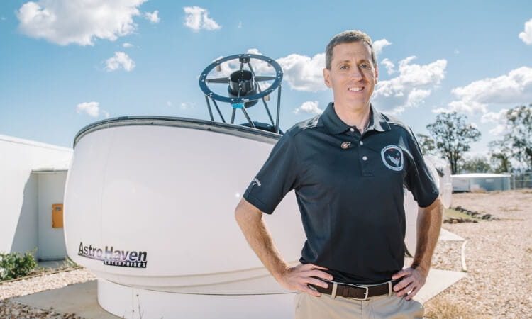 A man in a black polo shirt stands in front of a white observatory dome labeled "AstroHaven" under a clear blue sky.