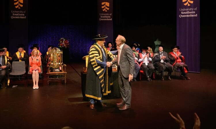 A ceremony at University of Southern Queensland with officials shaking hands on stage, surrounded by seated attendees in academic regalia.