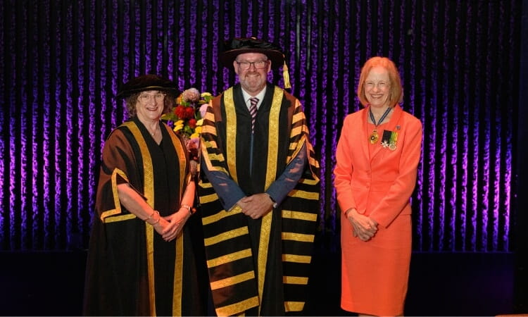 Three people in academic regalia and a formal suit stand in front of a purple curtain with flowers visible behind them.