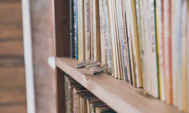 A wooden shelf filled with books, partially showing their spines. A pair of reading glasses rests on the shelf in front of the books.